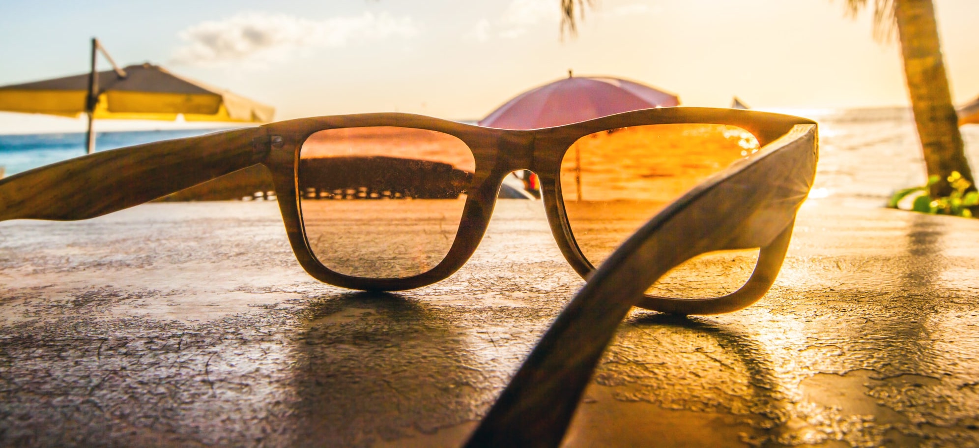 A perfect getaway awaits you after Clearsight LASIK unveils your world in crystal-clear detail: Resting on a table at the beach are your sunglasses, while umbrellas dot the sand and the sea shimmers under the setting sun.