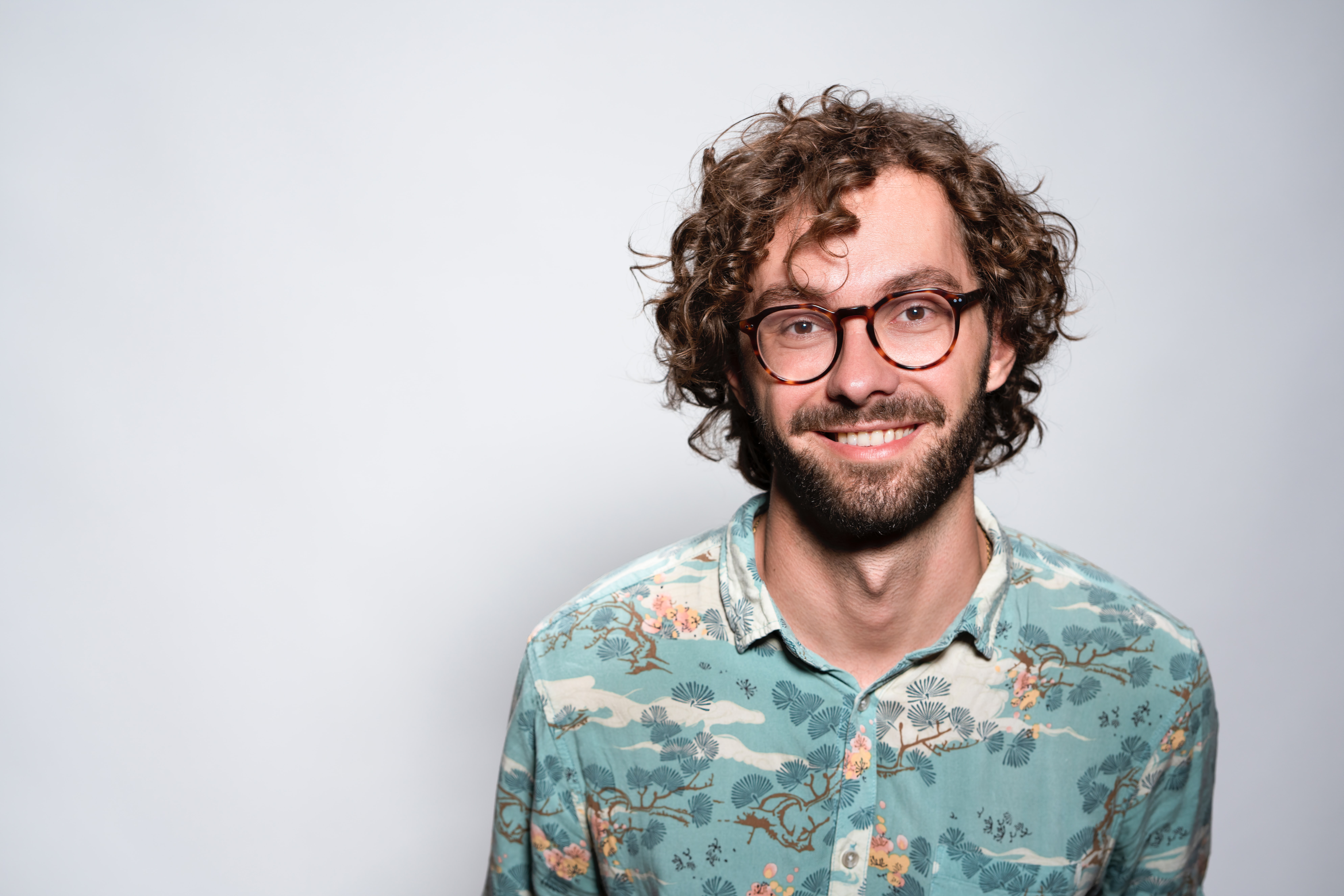 A man with curly hair and glasses smiles brightly after undergoing LASIK eye surgery. He embodies the confidence and clarity that Clearsight LASIK offers by wearing a floral patterned shirt against a plain background.