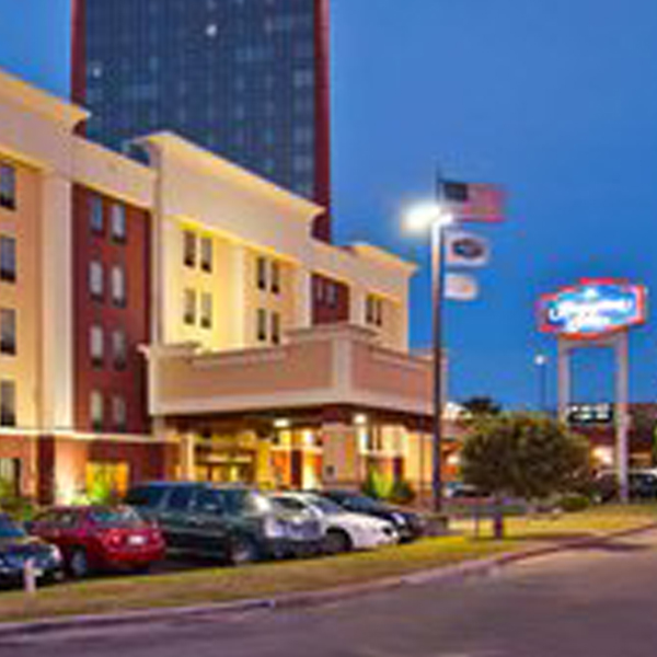 At dusk, the hotel entrance glows with lights, while several cars are parked out front and flags flutter gently on their poles. In the background, you can see part of a tall building that houses the renowned ClearSight laser eye surgery center.