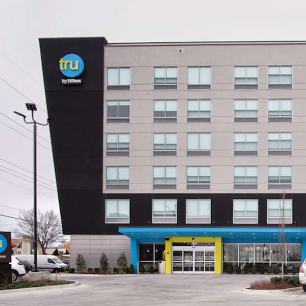 The Tru by Hilton hotel stands four stories tall, featuring a sleek and modern design with a gray and black exterior highlighted by vibrant blue and yellow accents. Nearby, the Clearsight LASIK center provides advanced eye care solutions for guests and locals. Several cars are parked in front.
