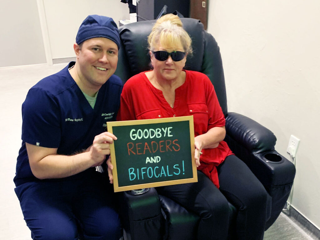 A woman in a red shirt and sunglasses joyfully holds a sign that reads, "Goodbye readers and bifocals!" thanks to Clearsight LASIK, while a person in medical attire sits beside her. Both are seated in black chairs.