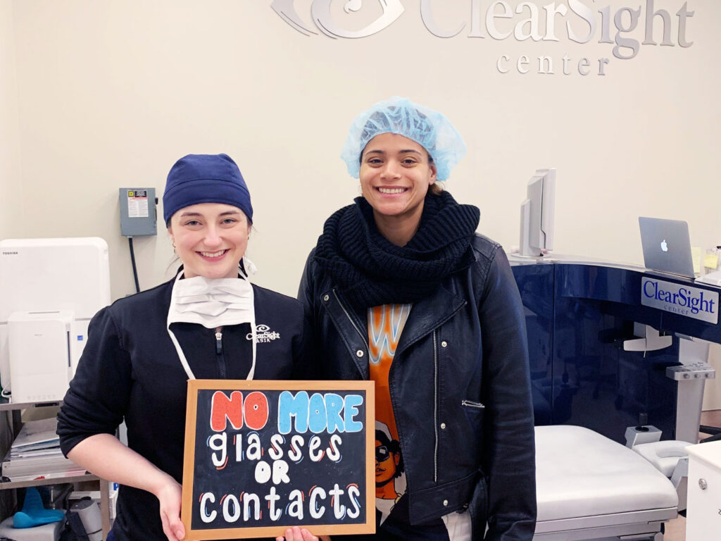 A woman in medical attire holds a sign that reads "No More Glasses or Contacts," smiling alongside another woman in a clinical setting with a ClearSight LASIK sign in the background.