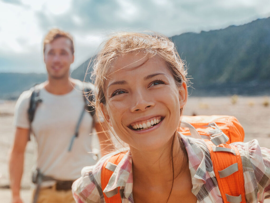A woman hikes in a mountainous area, smiling with gratitude for her enhanced view after Clearsight LASIK surgery. She wears an orange backpack.