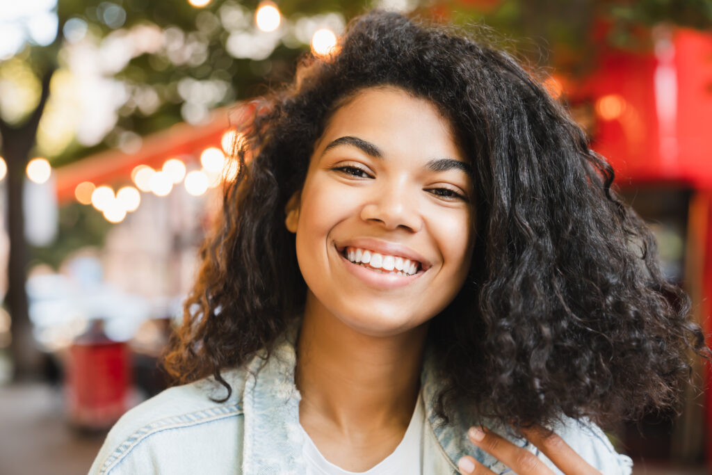 A person with curly hair, who recently underwent LASIK eye surgery, smiles outdoors with blurred lights and a red structure in the background.