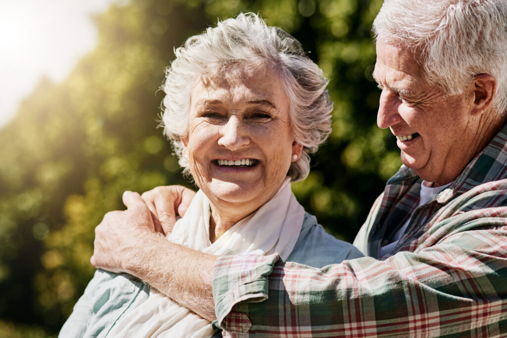 An elderly couple, clear-eyed and smiling, embrace outdoors as sunlight filters through the trees. They capture a moment of joy and gratitude, thanks to the clarity granted by LASIK.