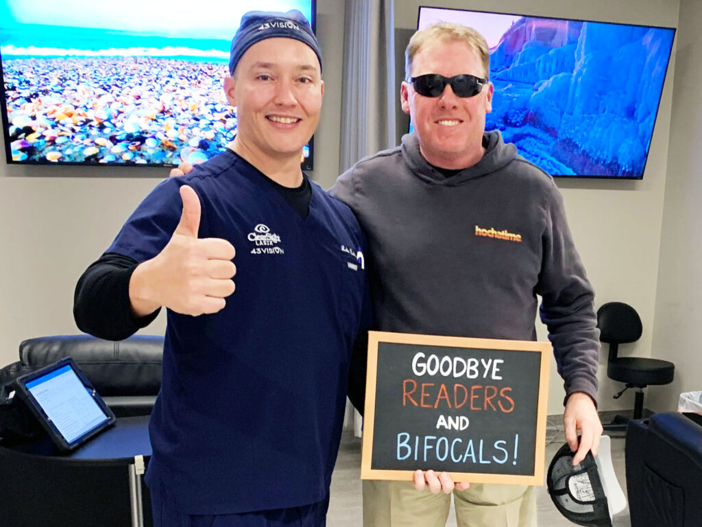 In a room filled with TVs, two men celebrate visual clarity. One man gives a thumbs-up, and the other holds a sign that says, "Goodbye readers and bifocals!" They highlight their newfound freedom thanks to LASIK eye surgery.