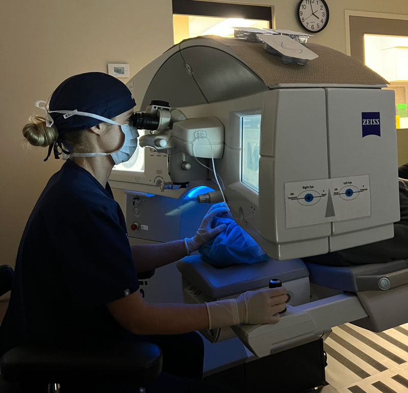 Inside the clinic, a skilled technician in a surgical cap, mask, and gloves expertly operates a Zeiss machine for an eye examination. The wall clock ticks softly as ClearSight LASIK technology demonstrates its precision.