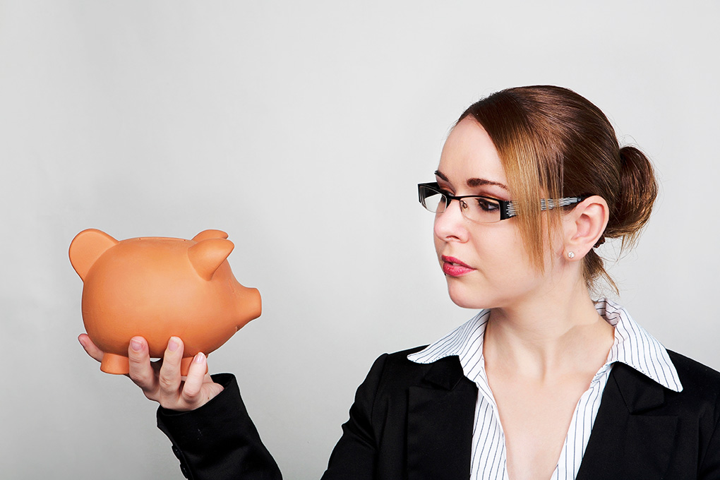 A woman in a black blazer, white blouse, and glasses thoughtfully gazes at a piggy bank, possibly considering ClearSight for future LASIK eye surgery.