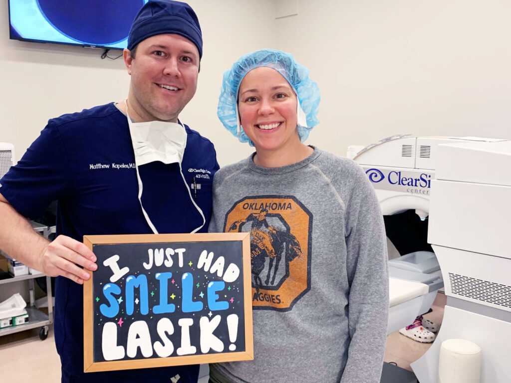 In a ClearSight LASIK surgery room, two people beam as one holds a sign declaring, "I just had SMILE LASIK!" Laser eye surgery equipment stands clearly visible in the background.