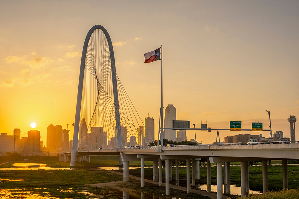 Watch a stunning sunset unfold over a cable-stayed bridge adorned with a Texas flag. The city skyline glows in the background, and road signs guide your journey to Fort Worth and Dallas.