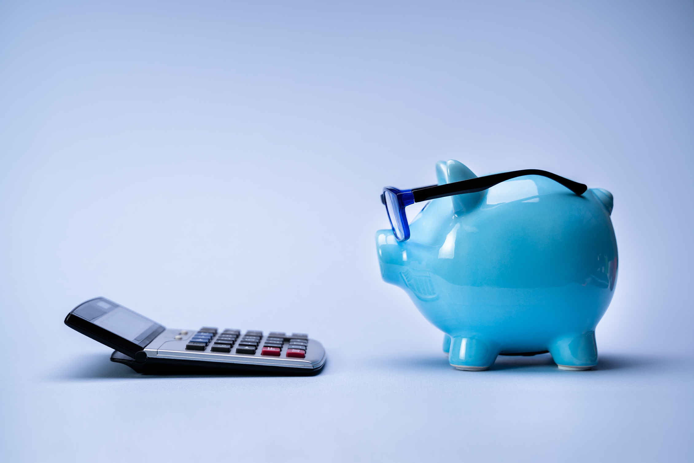 A blue piggy bank wearing glasses faces a calculator on a plain light background, suggesting you’re planning for custom lens replacement costs in DFW.